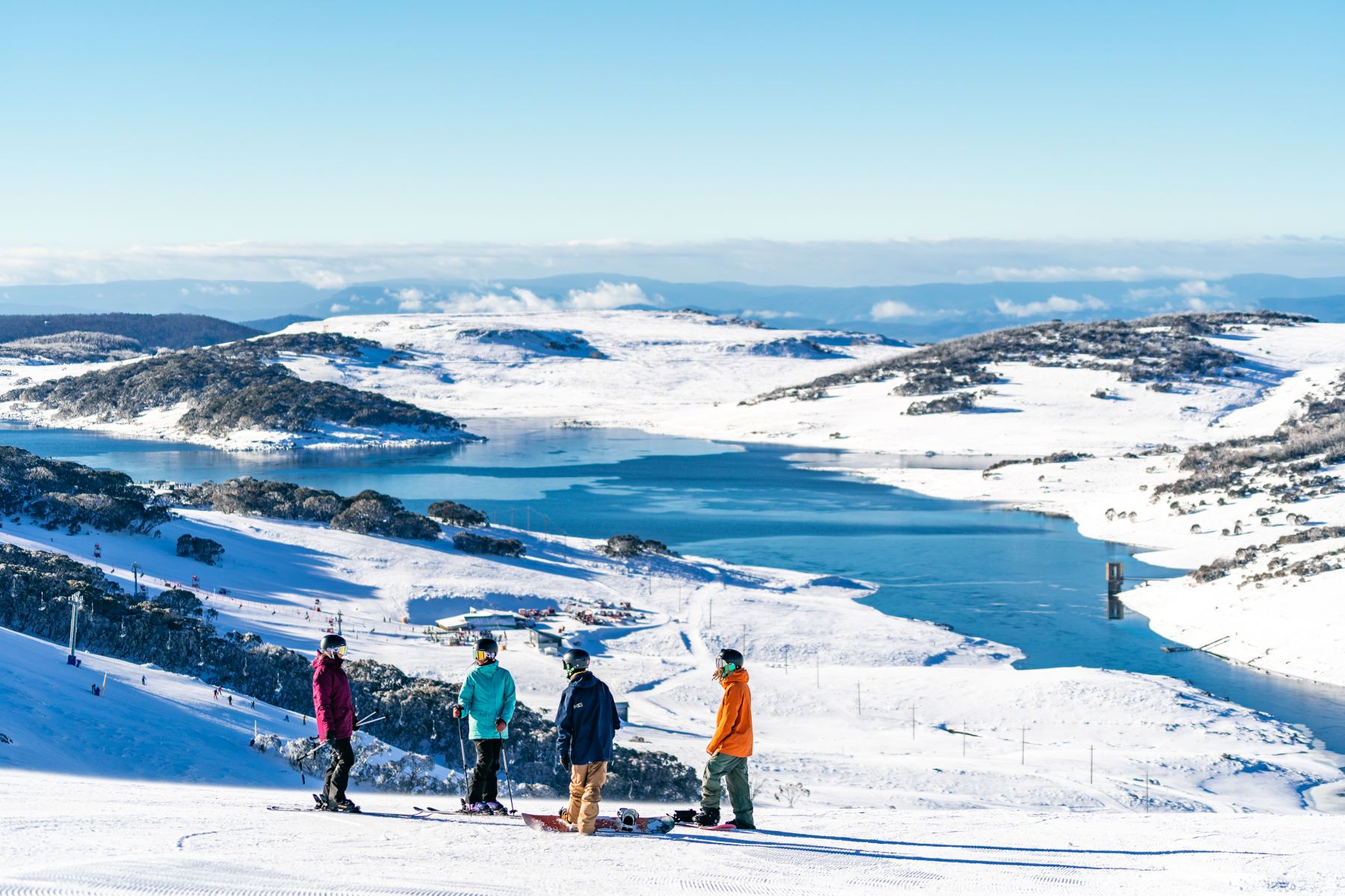 Two skiers and two snowboarders look over snow covered slopes and Falls Creek lake 